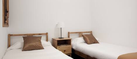 A room with two single beds, each with a wooden headboard, a bedside table with a lamp, and a small drawer.