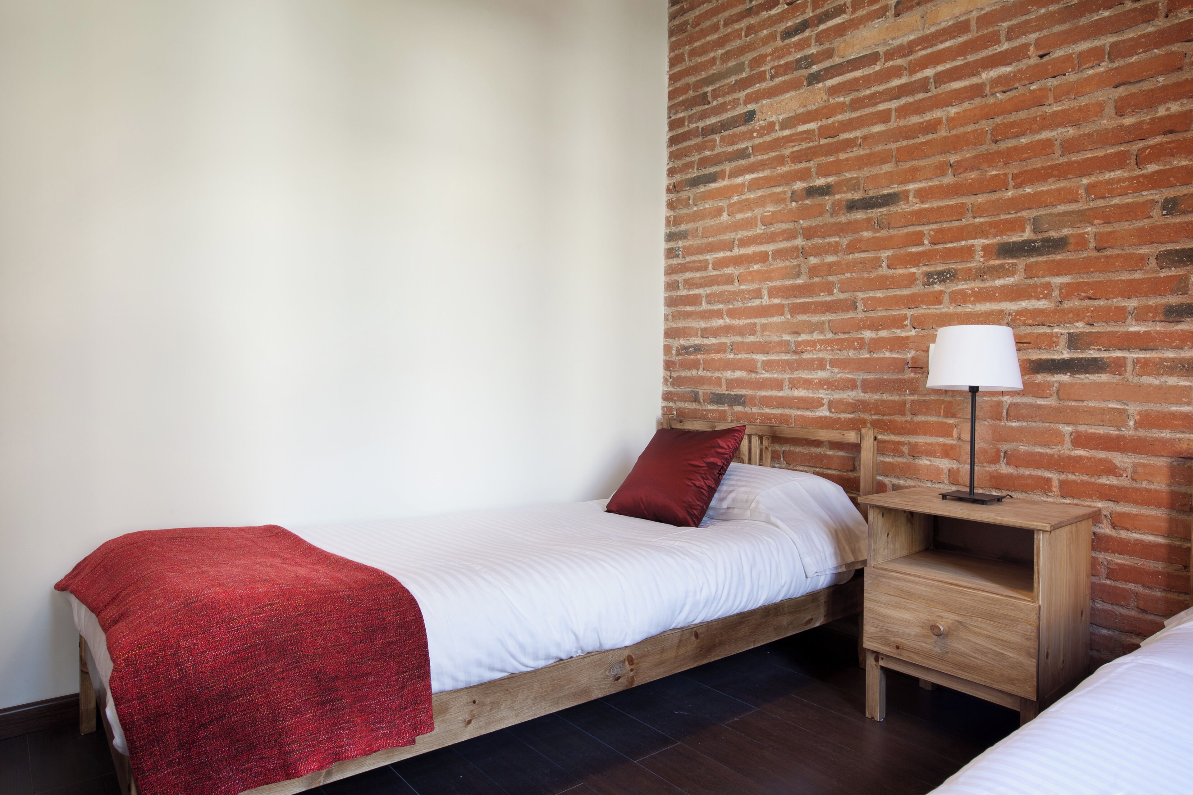 Un lit simple avec une couverture et des oreillers rouges, une table de chevet en bois avec une lampe, et un mur en briques.