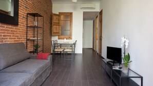 Ein modernes Wohnzimmer mit grauem Sofa, einem kleinen Esstisch mit Stühlen, einem Fernseher auf einem Schrank und einer Topfpflanze.