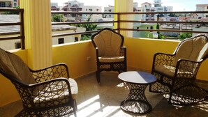 Een balkon met gele muren en uitzicht op gebouwen. Drie rieten stoelen en een kleine ronde tafel zijn aanwezig.
