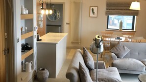 Ein modernes Wohnzimmer mit einer Couch, einem Couchtisch und einem Fernseher. Es gibt eine Küchenzeile mit Kühlschrank und Spüle. Der Raum hat eine hohe Decke mit freiliegenden Balken.