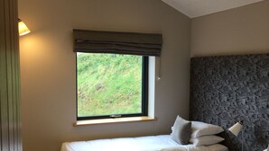 Ein Schlafzimmer mit zwei Betten, einem Fenster mit Blick auf Grünflächen, einem Nachttisch mit Lampe und einem strukturierten Kopfteil.