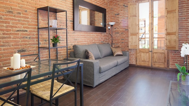 Een woonkamer met een bakstenen muur, een grijze bank, een glazen salontafel en een houten deur.