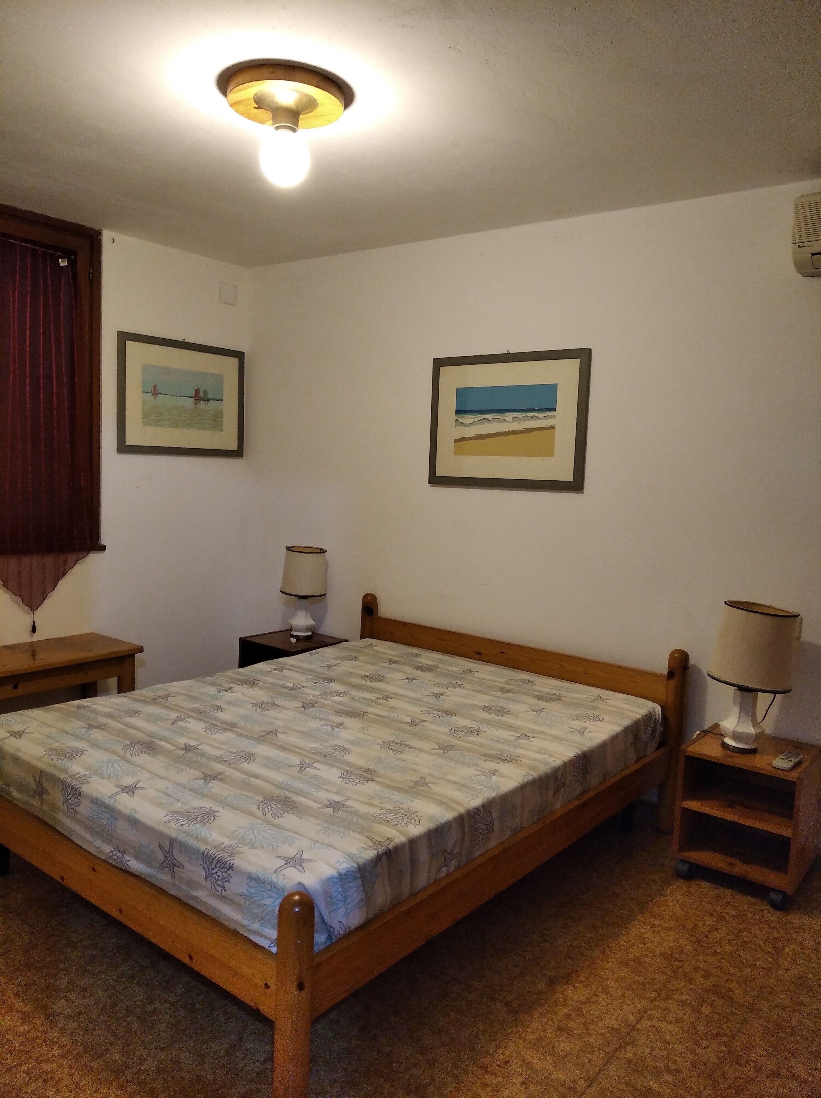 Un dormitorio con una cama de madera, dos mesitas de noche, una lámpara en el techo y dos cuadros en la pared.