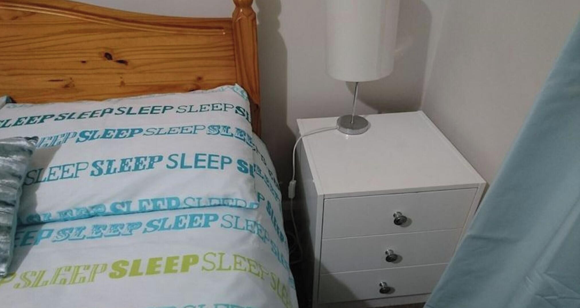 Une chambre à coucher avec une tête de lit en bois, une table de chevet blanche dotée de tiroirs et un lit recouvert d’une couette à motifs blancs et verts.