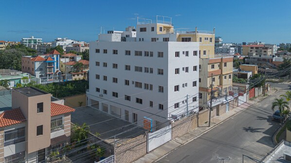 A multi-story building with a flat roof, surrounded by other buildings, a street, and some greenery.