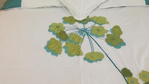 Una cama con colcha blanca y aguamarina, un plaid blanco de crochet y un estampado floral.