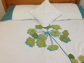 Una cama con colcha blanca y aguamarina, un plaid blanco de crochet y un estampado floral.