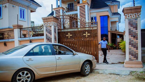 Un sedán color beige estacionado frente a la entrada porticada de una casa con puerta azul y una persona cerca.