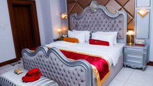 A bedroom with a tufted headboard, a bed with white and red bedding, a bedside table with a lamp, and a nightstand. - Room-limewood Hotel - Guest Room (Port Harcourt)