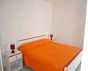 Ein Bett mit orangefarbener Bettdecke und weißem Kopfteil, zwei Nachttische mit roten Lampen sowie ein weißer Nachttisch mit Schublade.