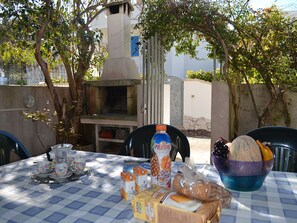 Uma área de refeições ao ar livre com uma mesa posta para dois, com toalha xadrez, uma garrafa de cerveja de Porto Rico, uma cesta de frutas e diversos alimentos.