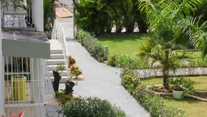 Ein Weg führt zu einem weißen Gebäude mit einem befestigten Eingangsbereich und einem kleinen Garten mit verschiedenen Pflanzen und Blumen.