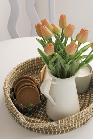 A white ceramic pitcher with tulips and a woven tray containing a ceramic mug and coasters.