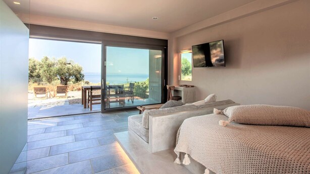 A modern bedroom with a large bed, a TV, and a balcony with a view of the sea. - Alterra Vita Eco Villas - Suite with Sea View (Neos Marmaras)