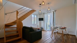 Ein moderner Wohnraum mit einer Holztreppe, einem schwarzen Sofa, einem Essbereich mit weißen Stühlen und einem an der Wand befestigten Fernseher.