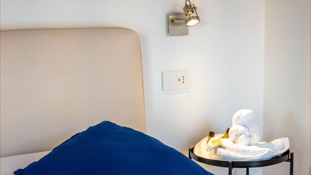 A neatly made bed with a blue pillow, a small side table with a towel, and a wall-mounted light fixture. - Appartamento Gondola Residence La Darsena (Lezzeno)