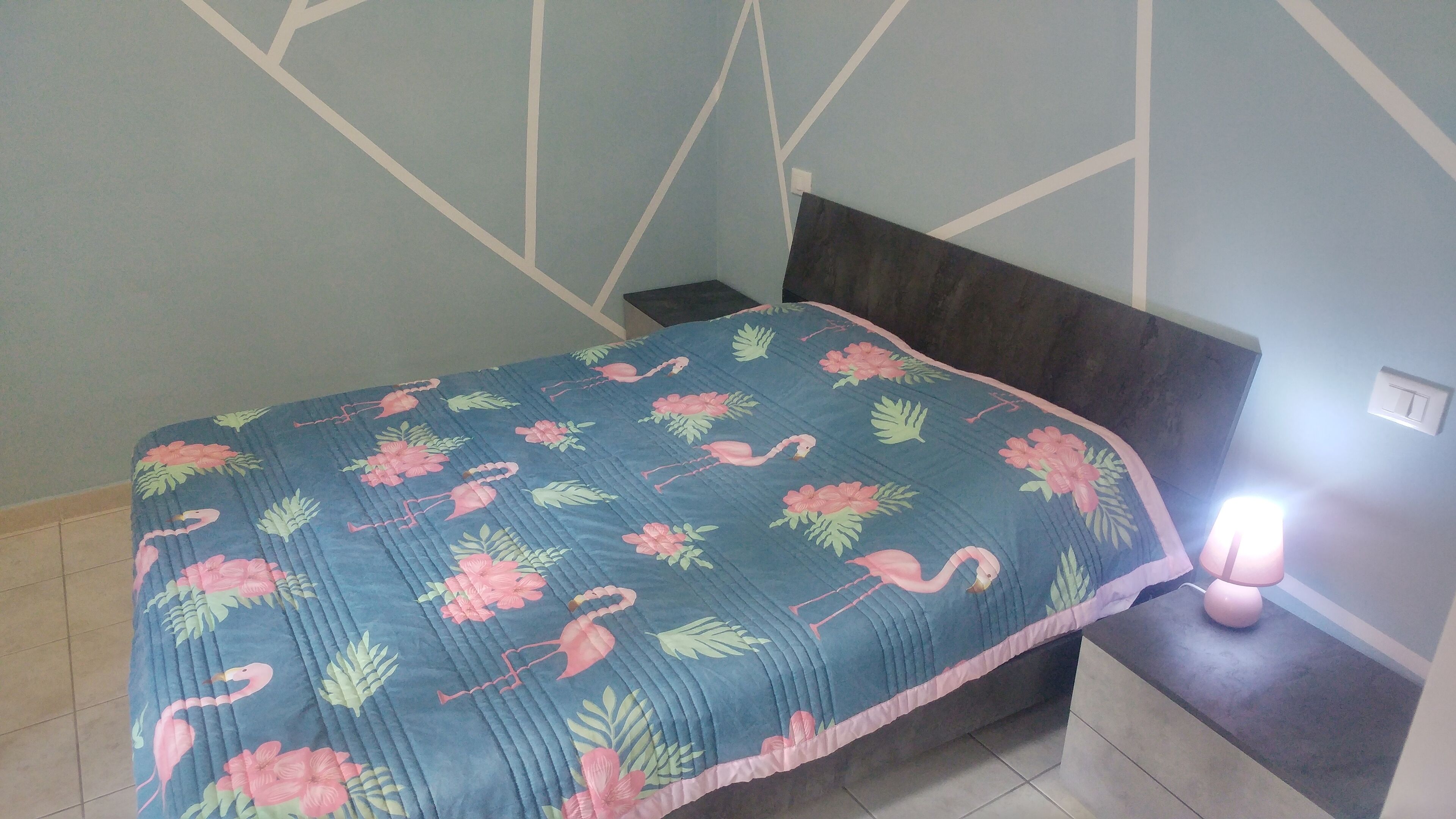 A bedroom with a bed covered by a floral-patterned quilt, a dark headboard, and a bedside lamp.