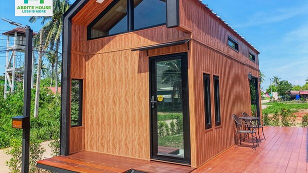 Ett modernt trähus med en veranda, stora fönster och en logotyp i hörnet.