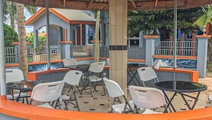 Un espace extérieur aménagé avec des chaises et des tables en plastique blanc, entouré d’un petit muret orange et d’une rambarde bleue.