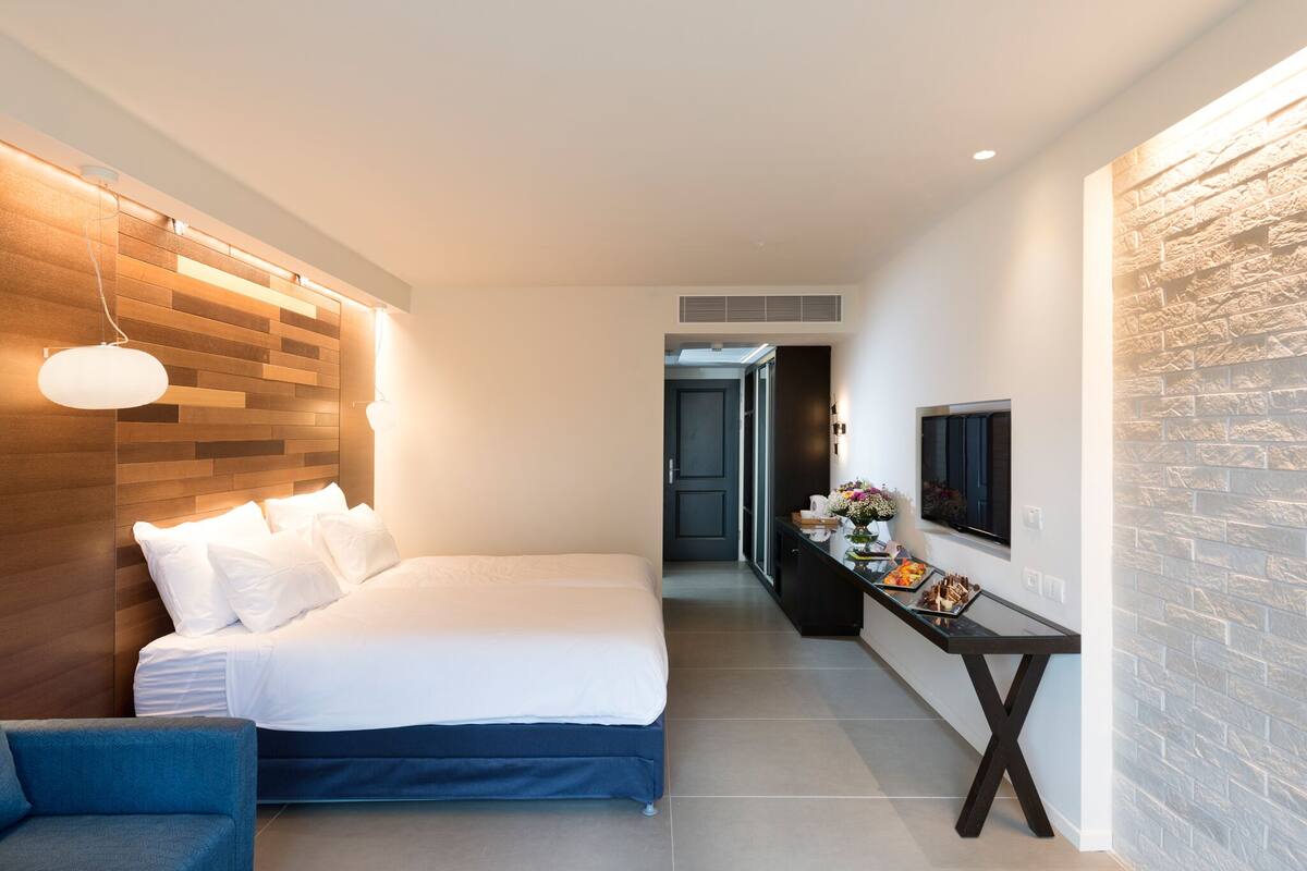 Quarto de hotel moderno com uma cama grande, um espaço de estar, uma pequena mesa com um vaso de flores e uma televisão montada.