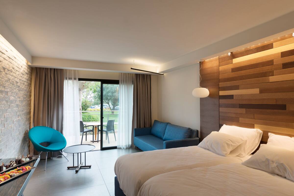 Quarto de hotel moderno com uma cama grande, um sofá, uma cadeira azul e um espaço de refeições com mesa e cadeiras.