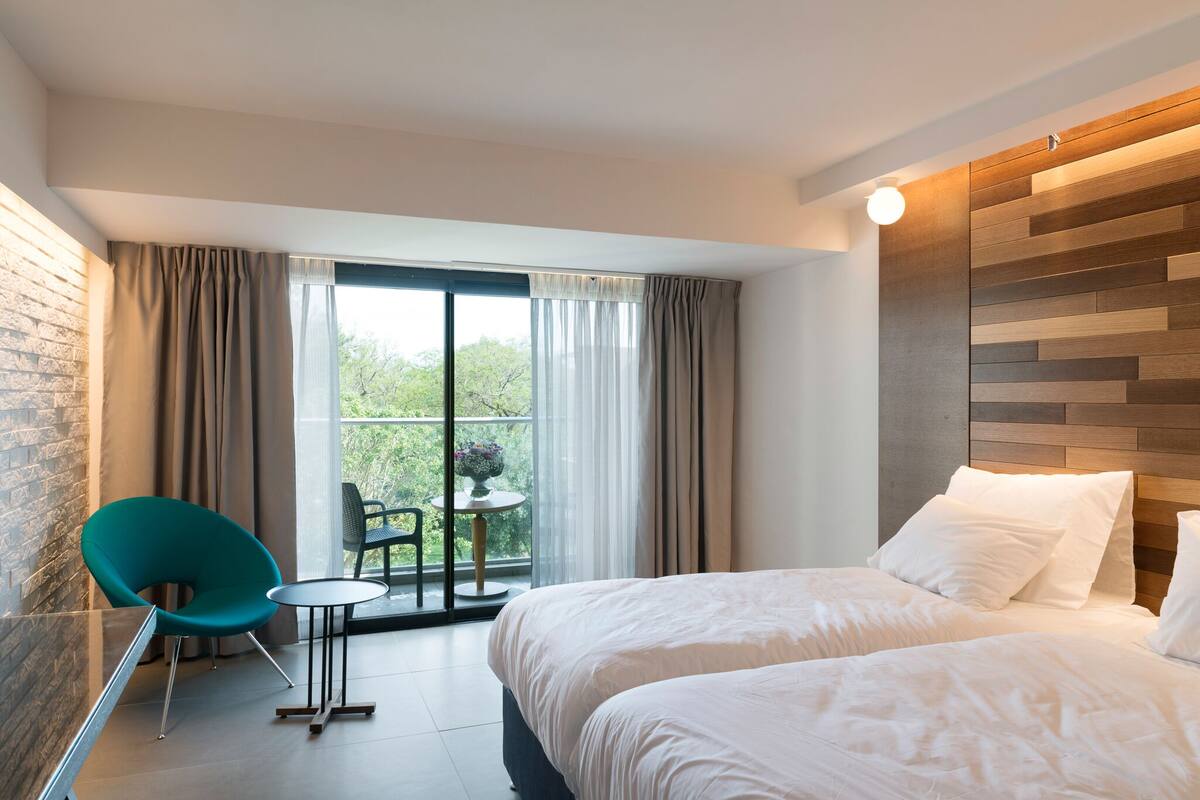 Quarto de hotel moderno com uma cama grande, uma mesinha, uma cadeira verde-água e uma varanda com vista para vegetação.