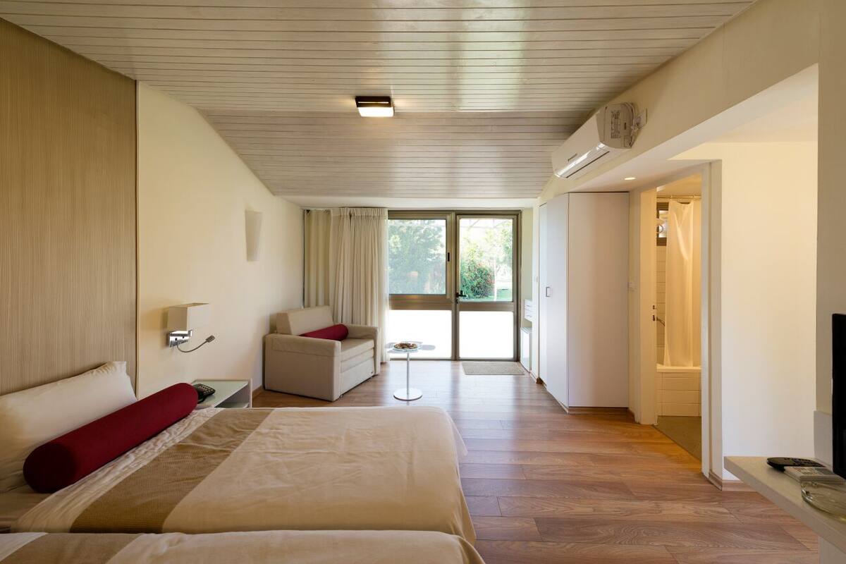 Quarto de hotel com uma cama grande, um sofá, uma mesa pequena e uma porta de vidro deslizante para uma varanda.