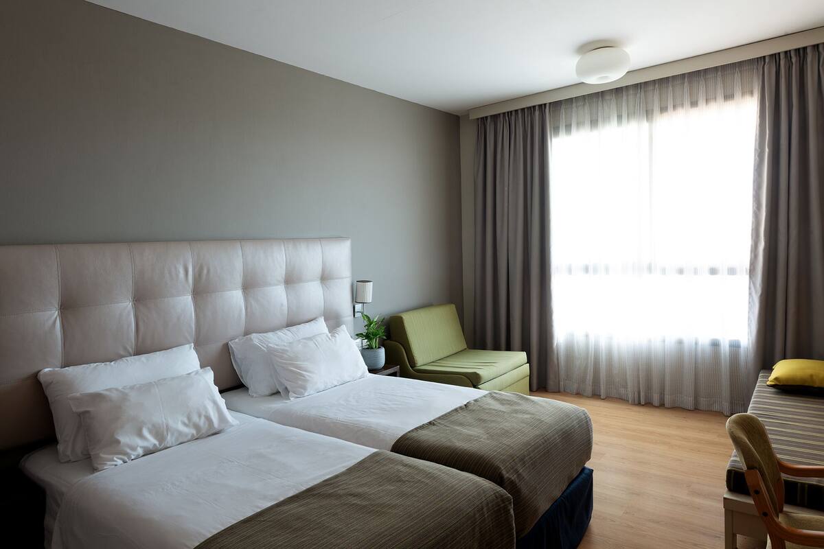 Quarto de hotel com duas camas, uma escrivaninha, uma cadeira e uma janela com cortinas.