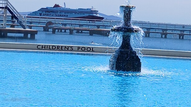 Una piscina infantil con una fuente en el centro, un crucero al fondo y un cielo azul despejado.