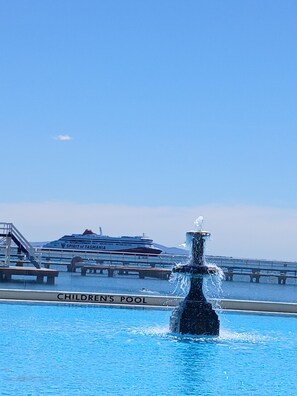 A children's pool with a fountain in the center, a cruise ship in the background, and a clear blue sky.