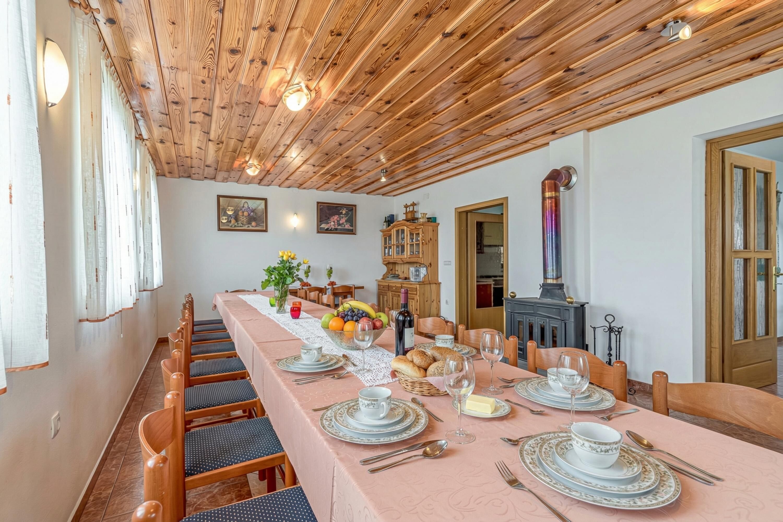 Une salle à manger avec une longue table dressée pour six personnes, des poutres en bois au plafond, une cheminée et un buffet aux portes vitrées.