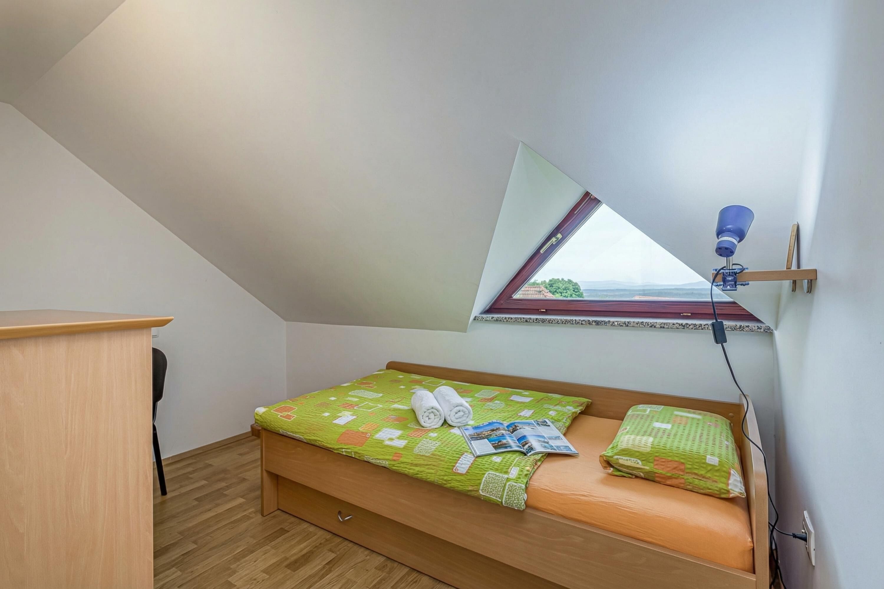 A small, single-bed room with a sloped ceiling, a wooden desk, a green bedspread, and a window with a view.