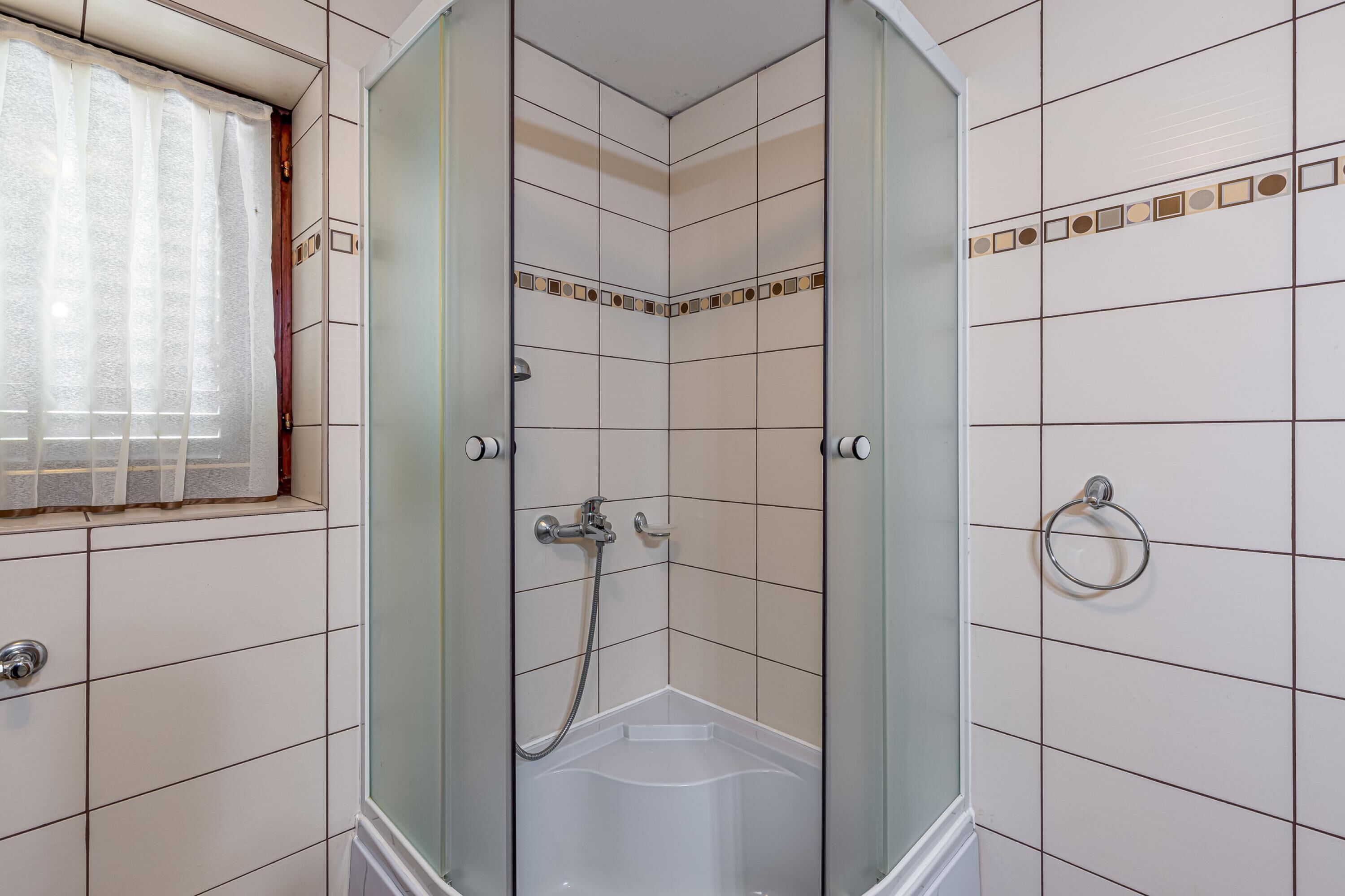 Eine Eckdusche mit Glastüren, ein Fenster mit leichtem Vorhang und ein Handtuchhalter an der Wand.