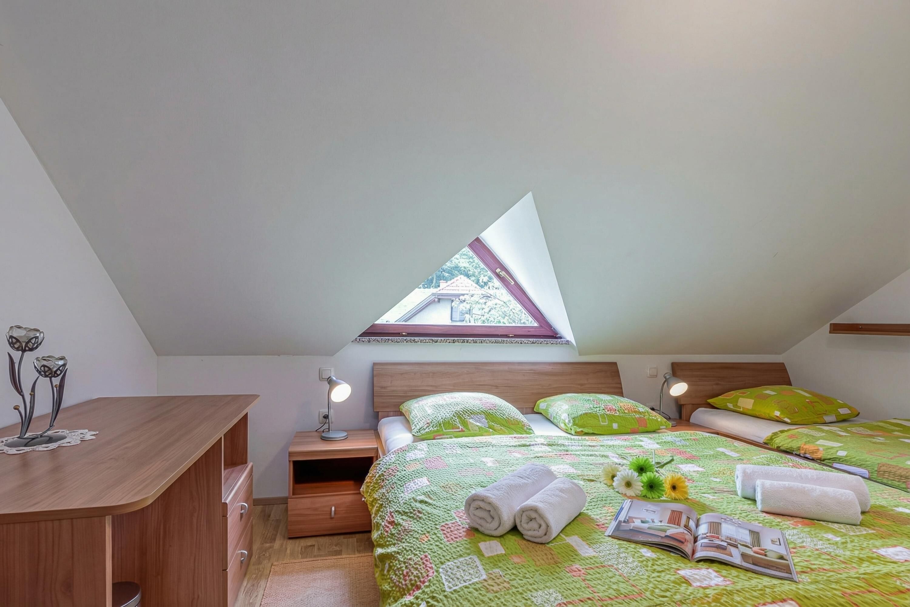A bedroom with a sloped ceiling, a wooden desk, a bedside table with a lamp, a bed with green bedding, and a skylight.