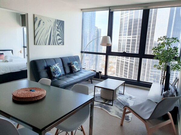 A modern living room with a sofa, coffee table, and chairs, large windows with a city view, and a dining area.