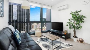 A modern living room with a black leather sofa, a coffee table, a flat-screen TV, a large window with a city view, and a potted plant.