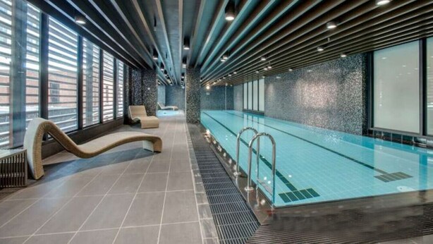 A modern indoor swimming pool with a long, narrow design, tiled flooring, and a seating area with lounge chairs.