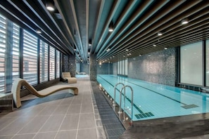 A modern indoor swimming pool with a long, narrow design, tiled flooring, and a seating area with lounge chairs.