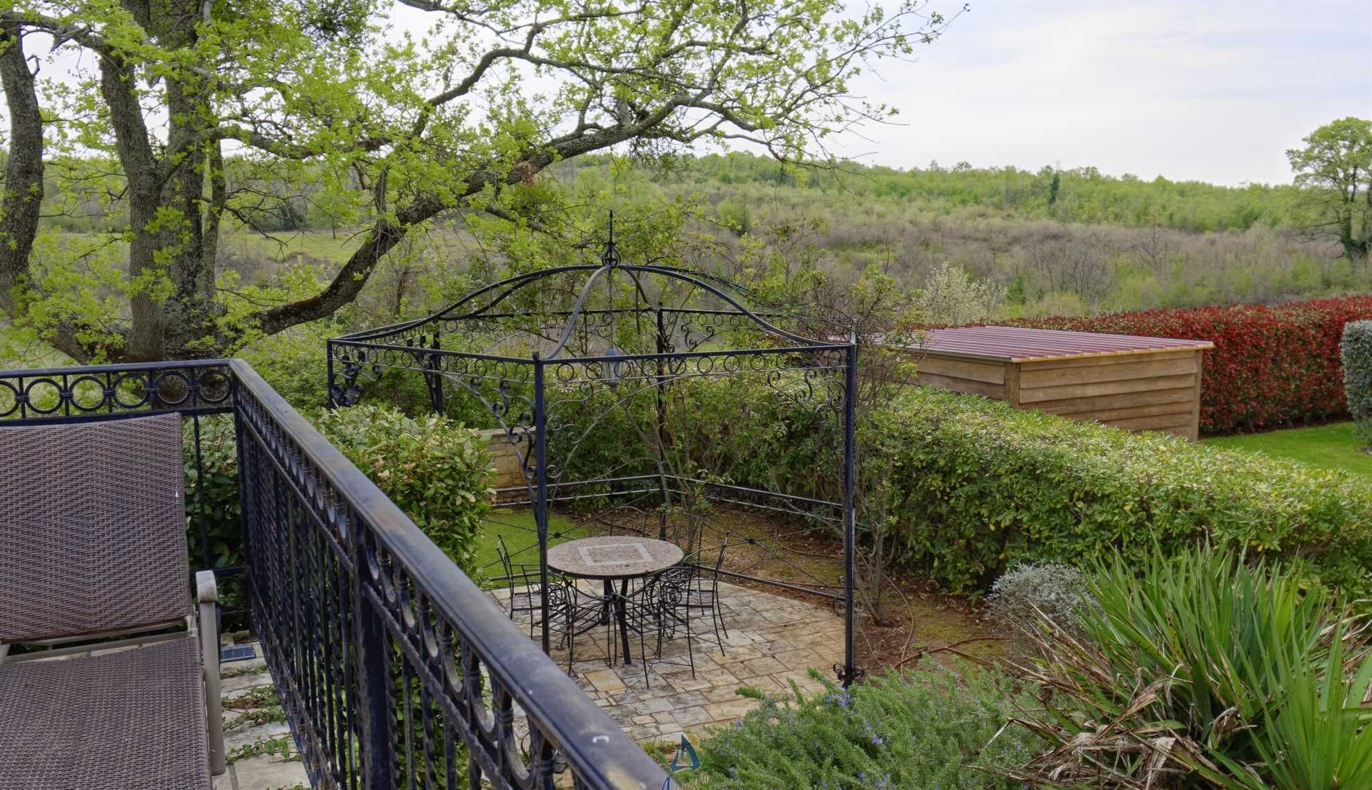 Une terrasse agrémentée d’un kiosque en métal, d’une remise en bois et d’une table en pierre avec des chaises.
