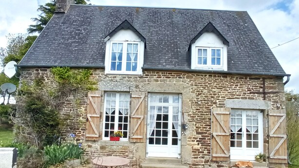 A stone house with two windows, a door, and a small patio with a table and chairs. - La Chaumière, Séjour Paisible (Sourdeval)