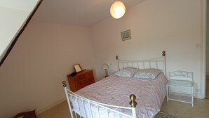 A bedroom with a white metal bed frame, a wooden dresser, a bedside table, and a hanging light fixture. - La Chaumière, Séjour Paisible (Sourdeval)