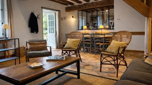 A cozy living area with a wooden coffee table, wicker chairs, a sofa, and a dining table with chairs. A kitchen area is visible in the background with a bar and counter.