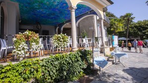 An outdoor café with a blue mural ceiling, white chairs, and tables.