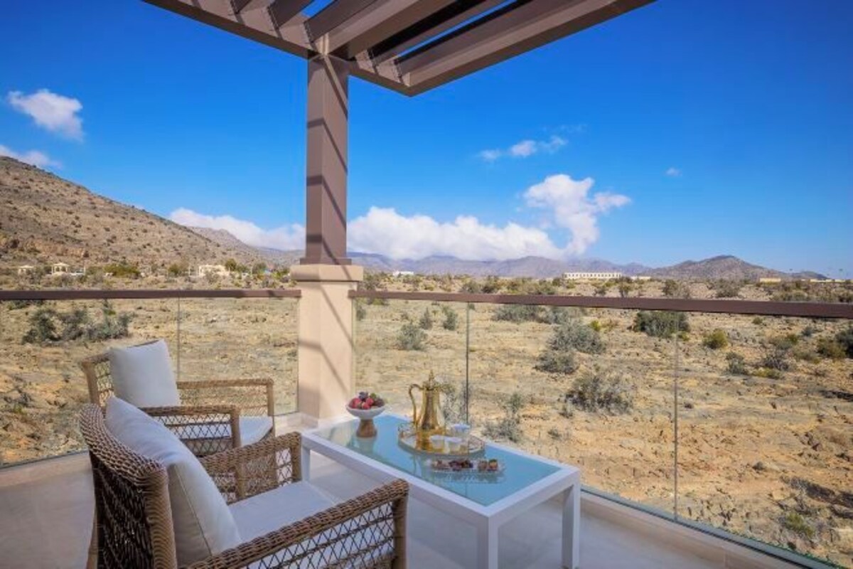 A balcony with wicker furniture and a glass table, overlooking a desert landscape with mountains in the distance.