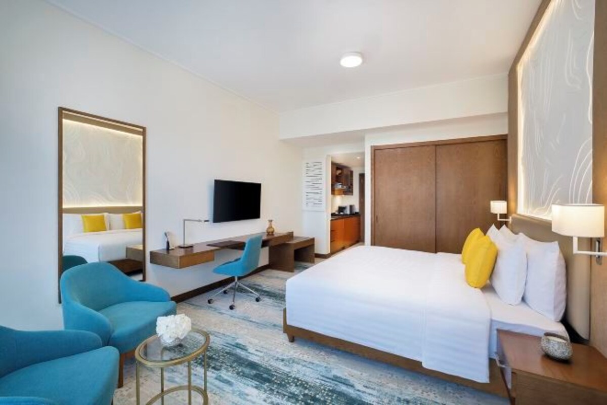 A hotel room with a bed, two blue armchairs, a desk with a chair, a TV, and a wardrobe.