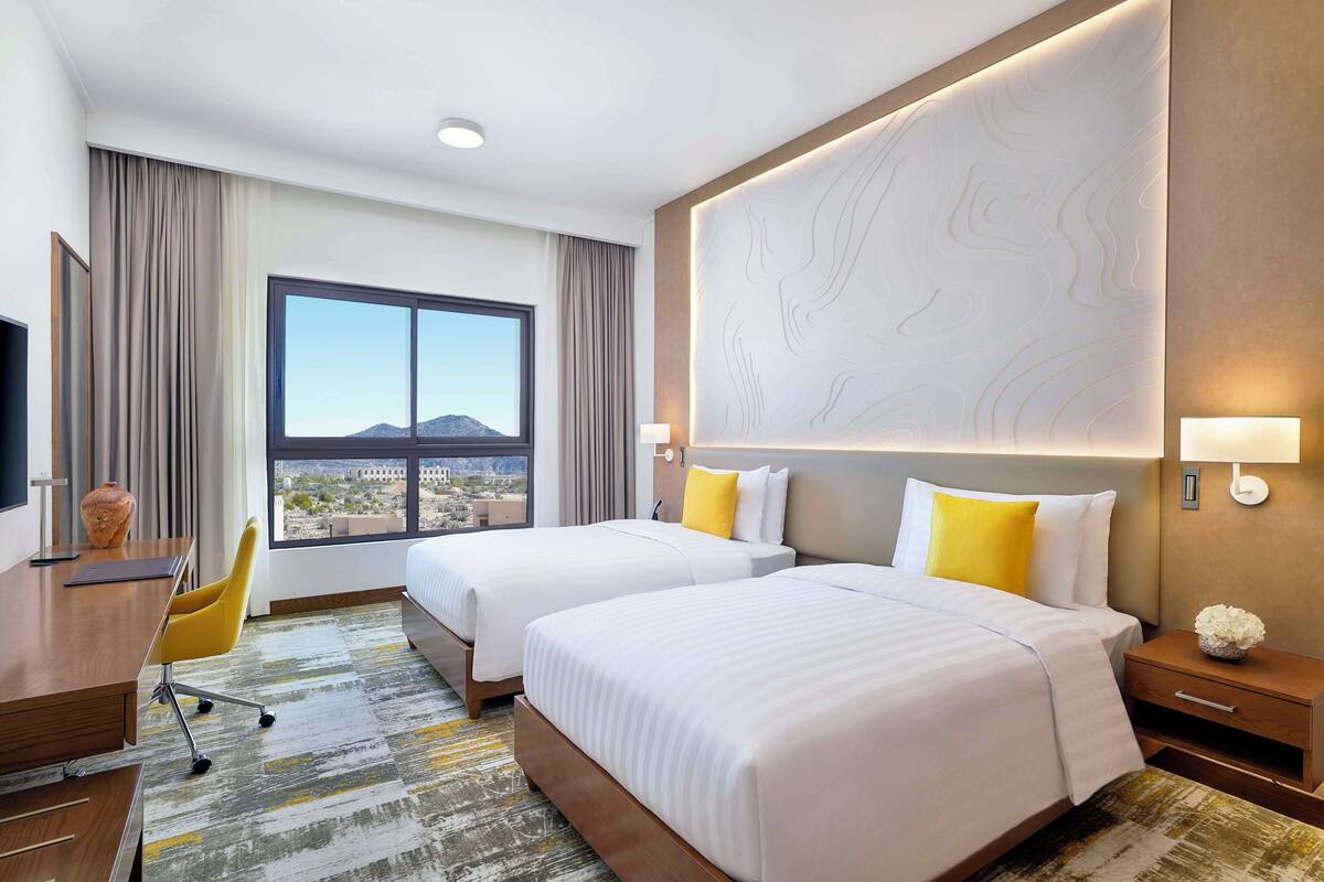 A hotel room with two beds, a desk, a chair, a large window with a view, and a decorative wall panel.