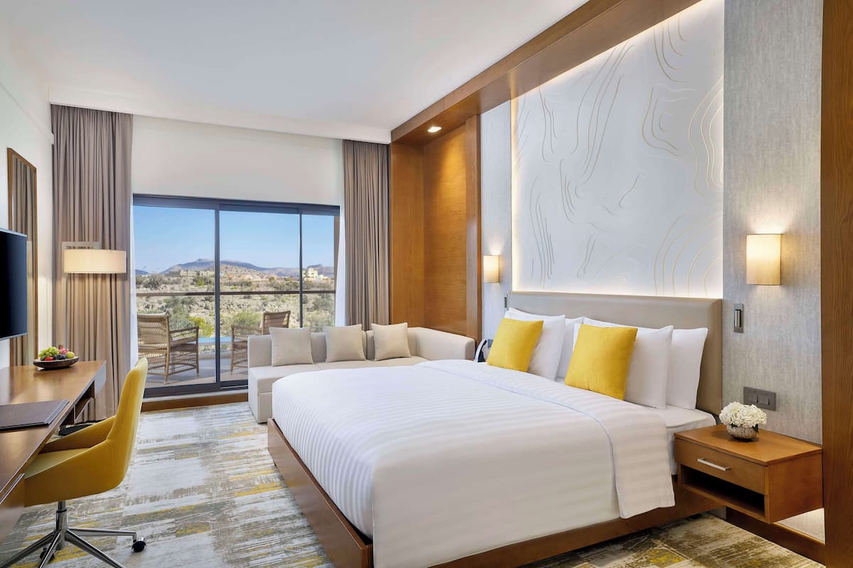A hotel room with a large bed, a desk with a chair, a TV, and a view of the city through large windows.