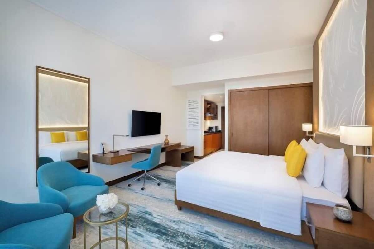 A hotel room with a bed, two blue armchairs, a desk with a chair, a TV, and a wardrobe.