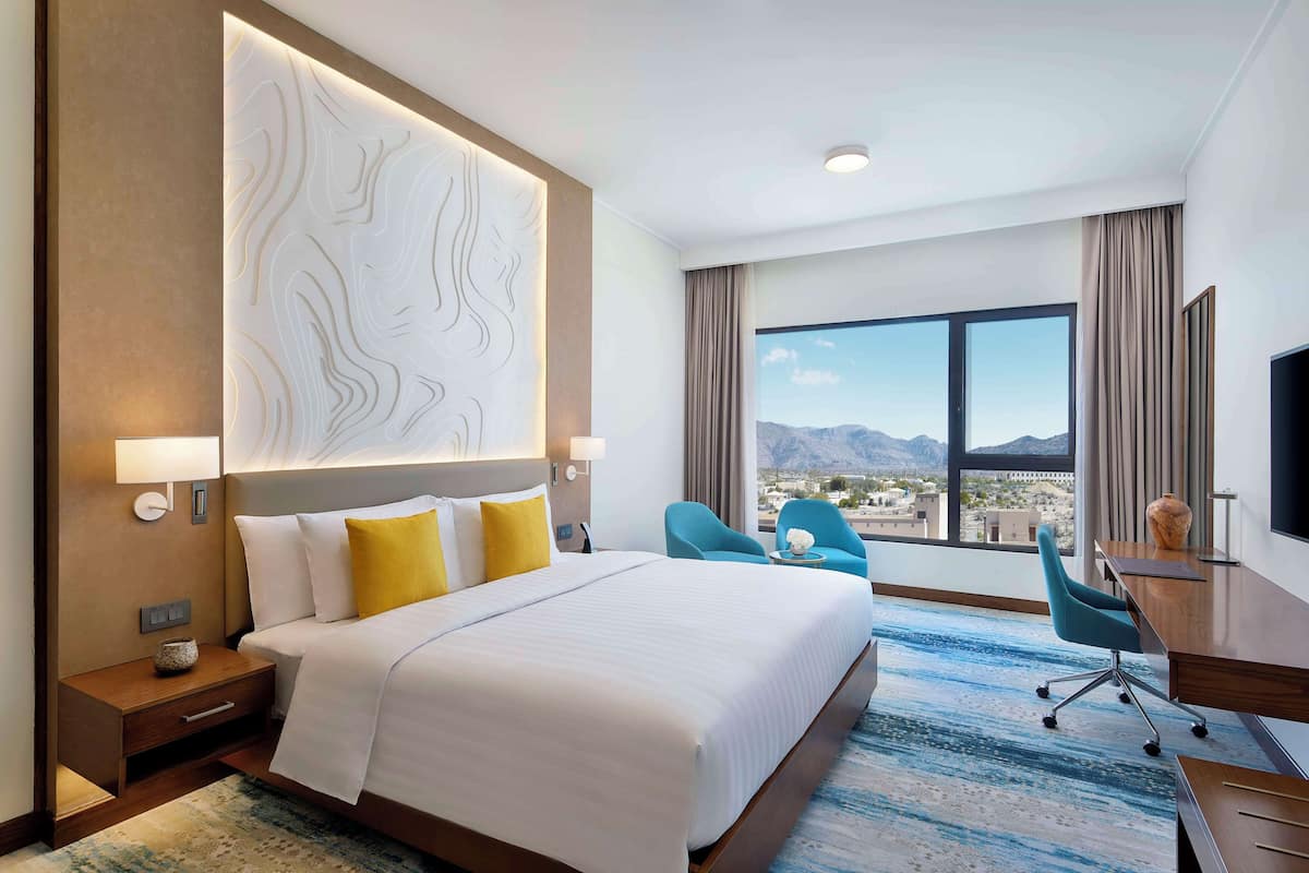 A hotel room with a large bed, two chairs, a desk, and a large window with a mountain view.
